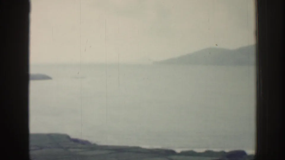 1961 Ireland coastal frame showing pale water, a dark foreground shoreline, and soft distant mountains.