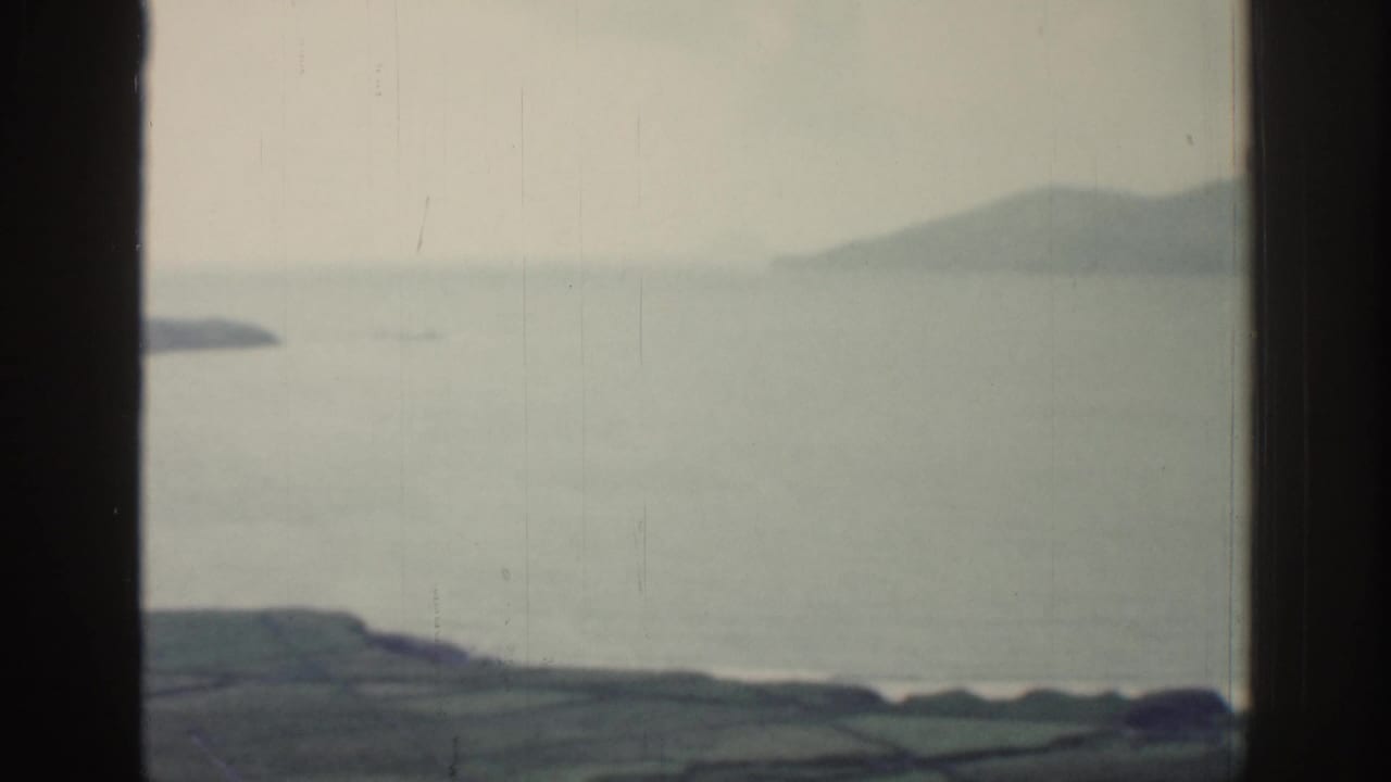 1961 Ireland home-movie frame showing water, distant hills, and patchwork fields seen through dark window mullions.