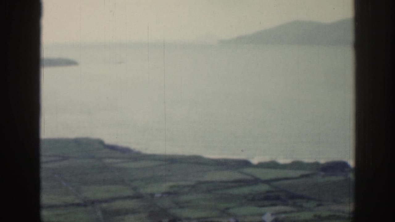 1961 Ireland countryside view with a dark window mullion, calm water, distant landforms, and divided green fields.