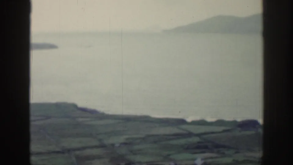 1961 Ireland countryside view with a dark window mullion, calm water, distant landforms, and divided green fields.