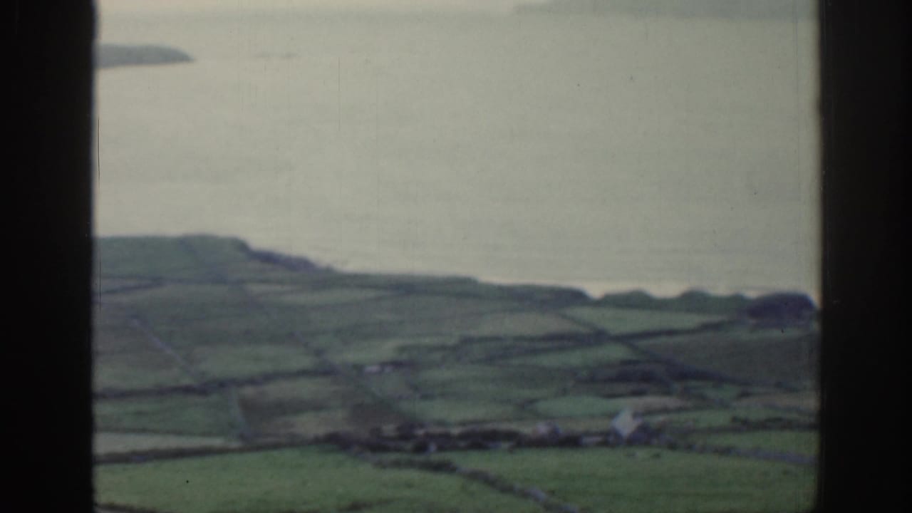 1961 Ireland frame looking downward toward green fields, a pale shoreline, and calm water beyond the window frame.