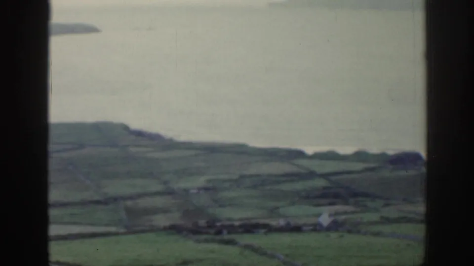 1961 Ireland frame looking downward toward green fields, a pale shoreline, and calm water beyond the window frame.