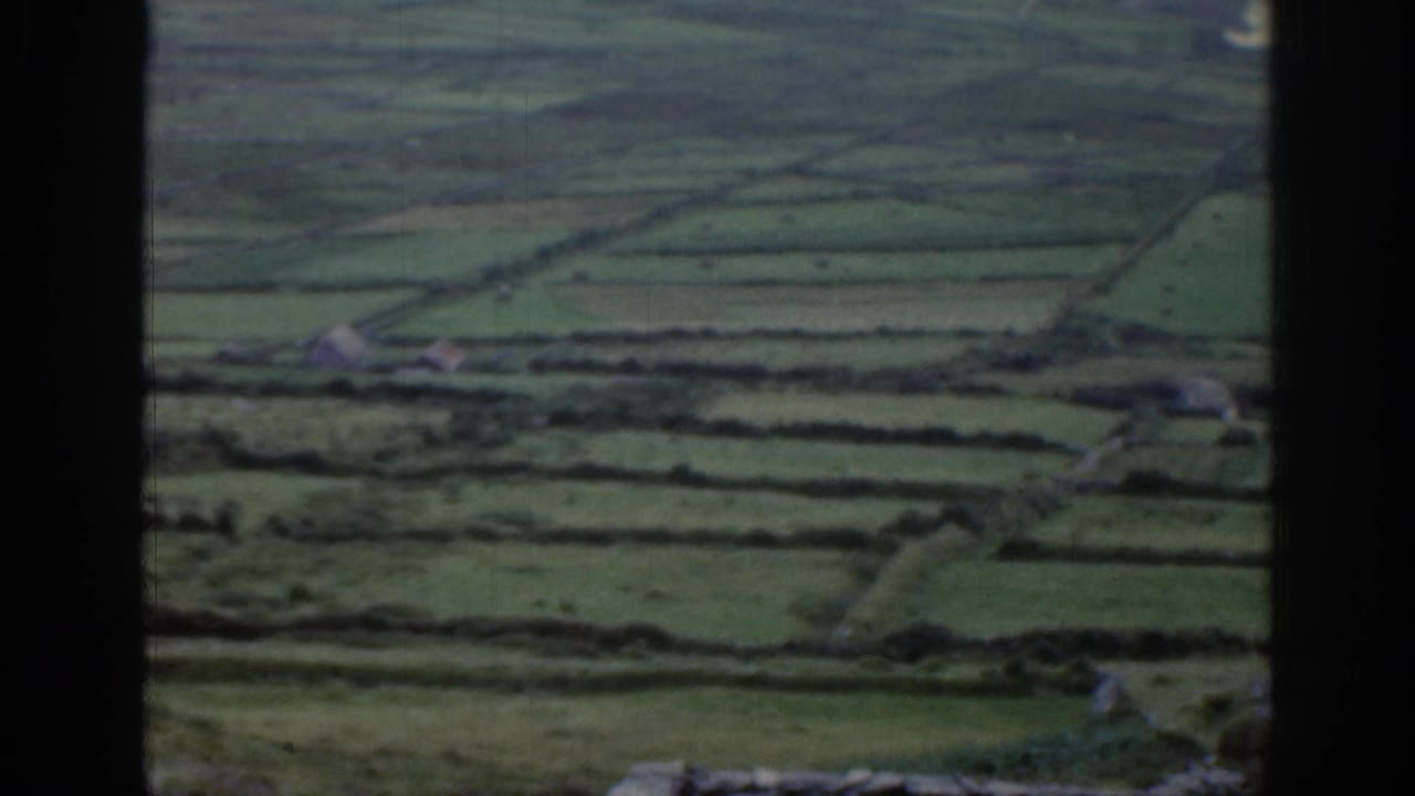 1961 Ireland home-movie frame focused on patchwork green fields, stone walls, hedgerows, roads, and scattered small buildings.