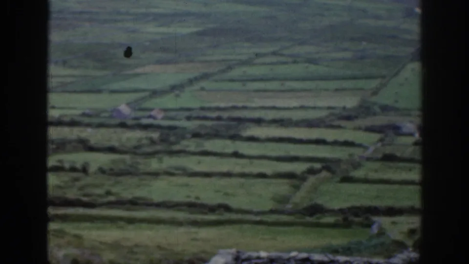 1961 Ireland home-movie view of green fields, stone wall boundaries, distant farm buildings, and a dark foreground frame.