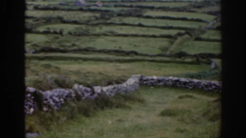 8mm view of rural Ireland showing grassy fields, low stone walls, foreground vegetation, and a dark window-frame edge.