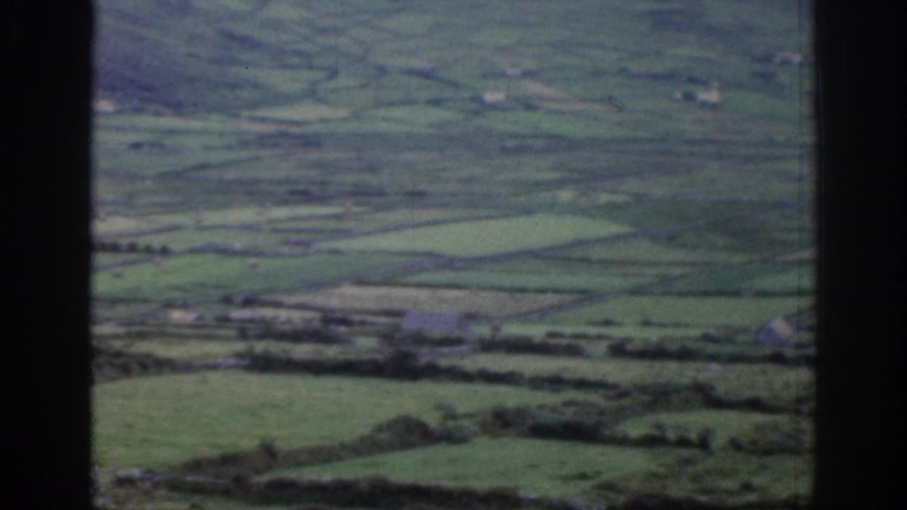 Wide rural Ireland landscape with divided green fields, stone or hedge boundaries, distant hills, and soft faded 8mm color.