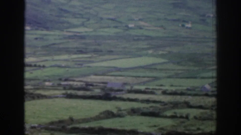 Wide rural Ireland landscape with divided green fields, stone or hedge boundaries, distant hills, and soft faded 8mm color.