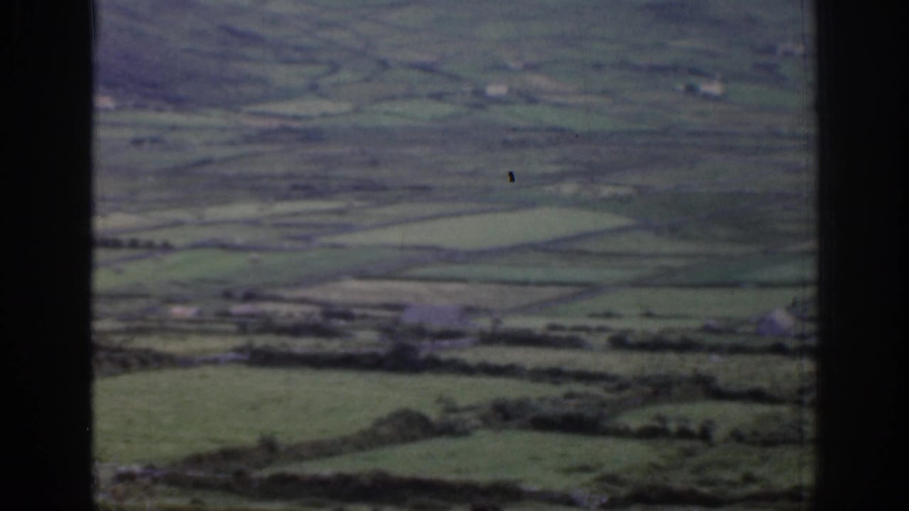 1961 Ireland home-movie frame looking across a green rural valley with patchwork fields, distant buildings, and dark frame borders.