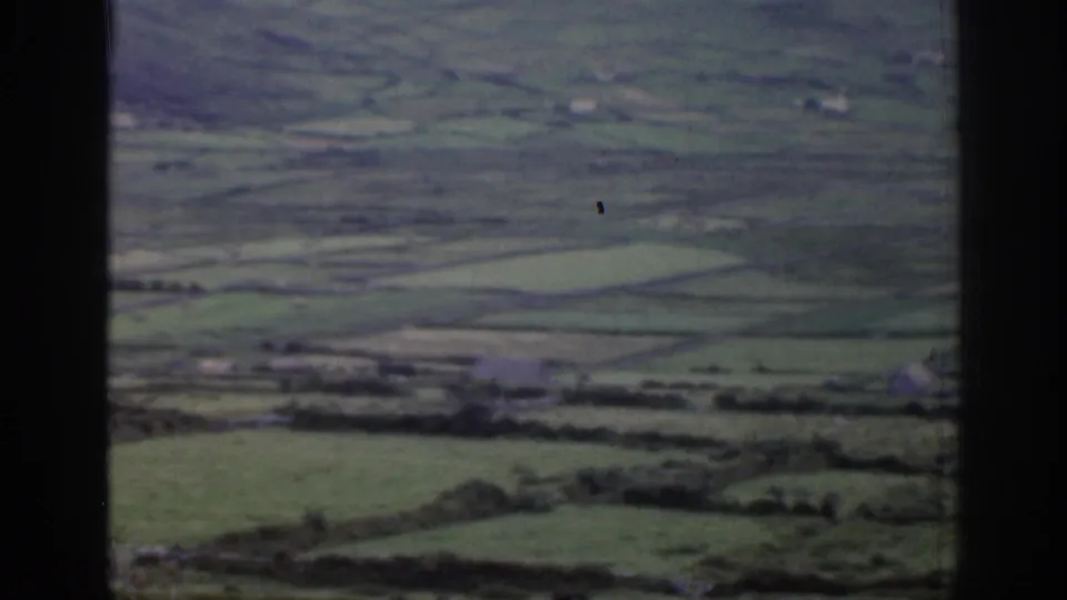 1961 Ireland home-movie frame looking across a green rural valley with patchwork fields, distant buildings, and dark frame borders.