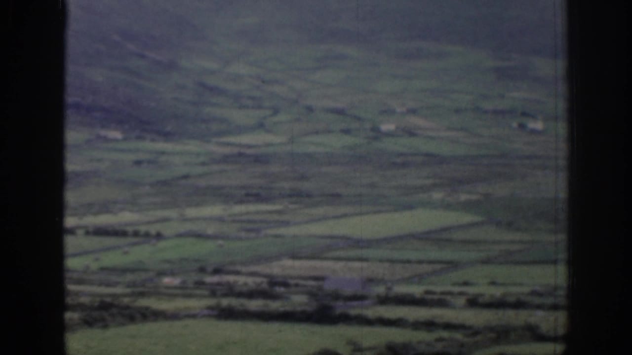 1961 Ireland 8mm frame showing a hazy rural valley, divided fields, distant farm buildings, and dark border areas from the filmed view.