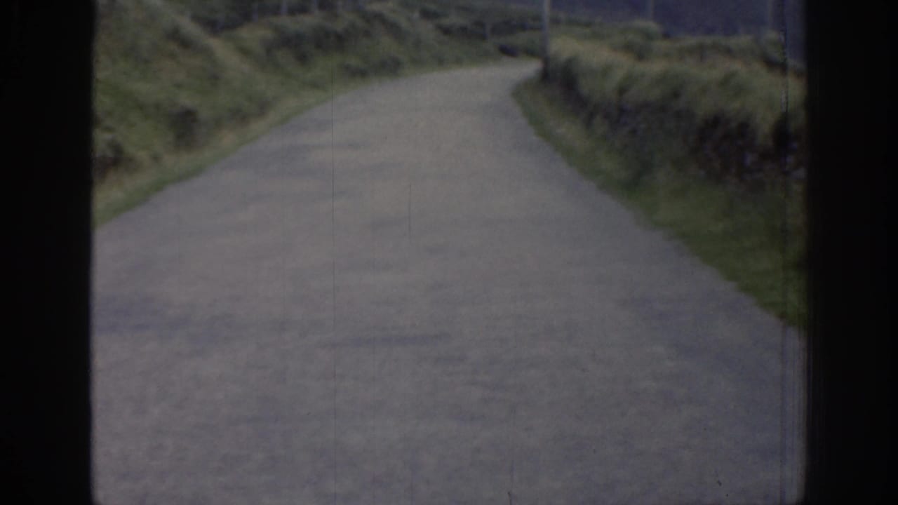 1961 Ireland home-movie frame of an empty grey country road bordered by overgrown grassy banks and muted rural hills beyond.