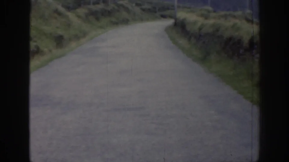1961 Ireland home-movie frame of an empty grey country road bordered by overgrown grassy banks and muted rural hills beyond.