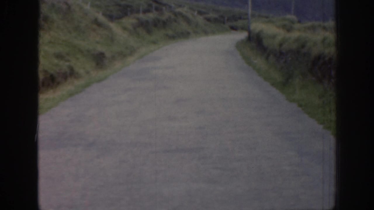 1961 Ireland 8mm frame showing a narrow rural road curving through grassy hillsides, seen from a moving vehicle.