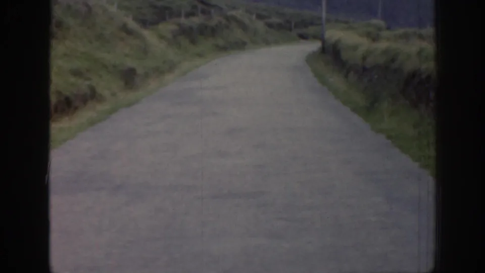 1961 Ireland 8mm frame showing a narrow rural road curving through grassy hillsides, seen from a moving vehicle.