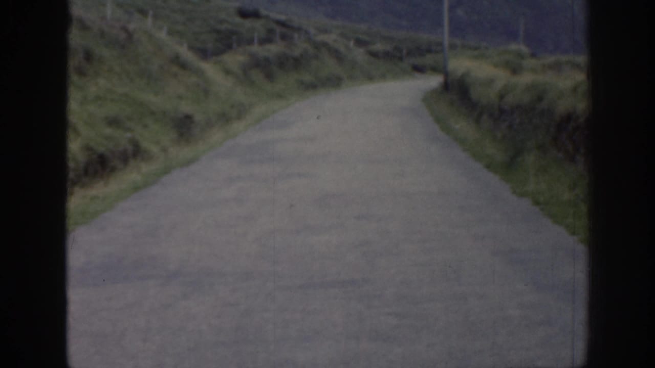 1961 Ireland home-movie frame with a winding rural road bordered by green banks, low walls, and roadside posts.