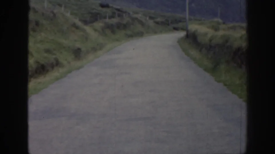 1961 Ireland home-movie frame with a winding rural road bordered by green banks, low walls, and roadside posts.