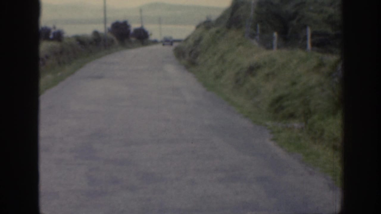 1961 Ireland 8mm frame showing the road extending ahead through grassy roadside banks toward a distant open area.