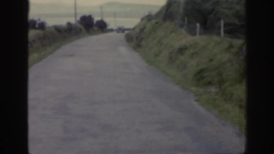1961 Ireland 8mm frame showing the road extending ahead through grassy roadside banks toward a distant open area.