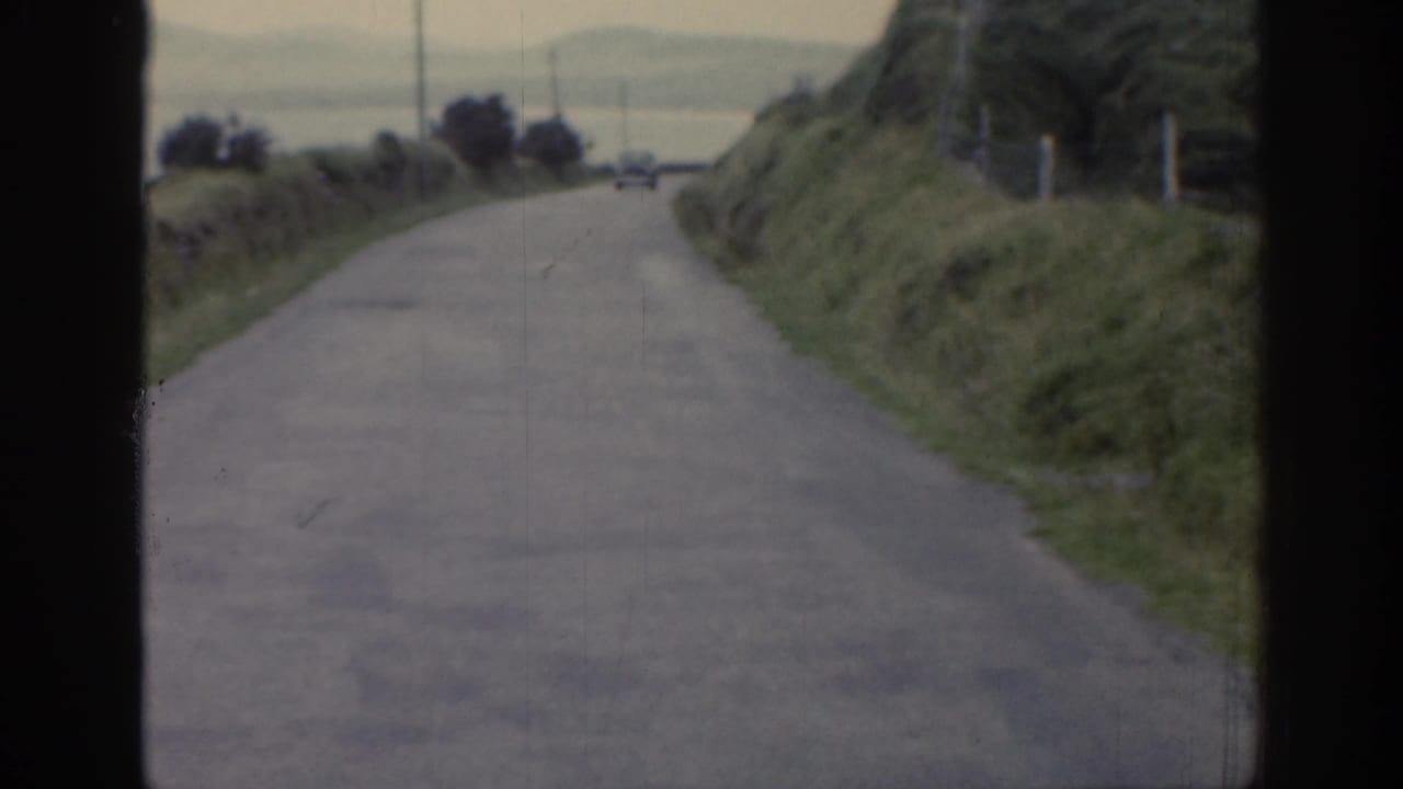 1961 Ireland 8mm frame from a moving vehicle, with a narrow road, grassy banks, poles, and a distant car ahead.