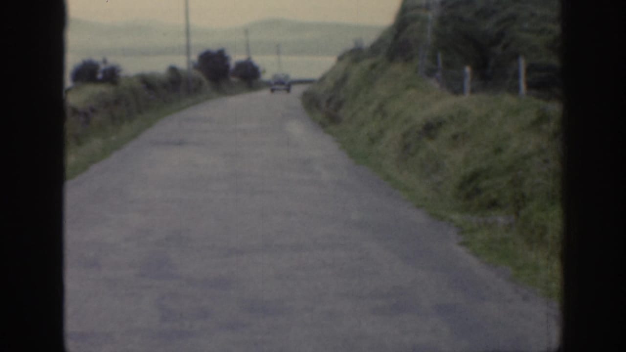 1961 Ireland home-movie frame of a narrow rural road running between green hillsides with a small distant car visible ahead.