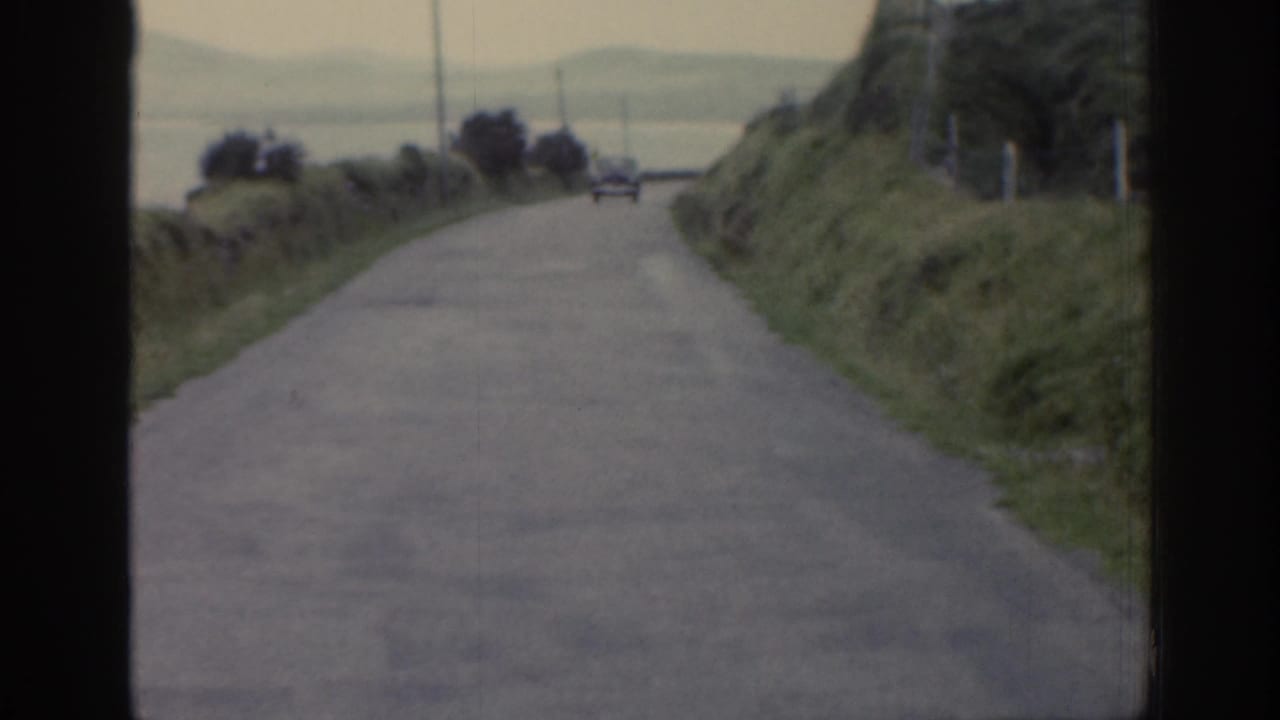 1961 Ireland countryside view from a moving vehicle, showing a winding road, grassy slopes, roadside edges, and a distant car.