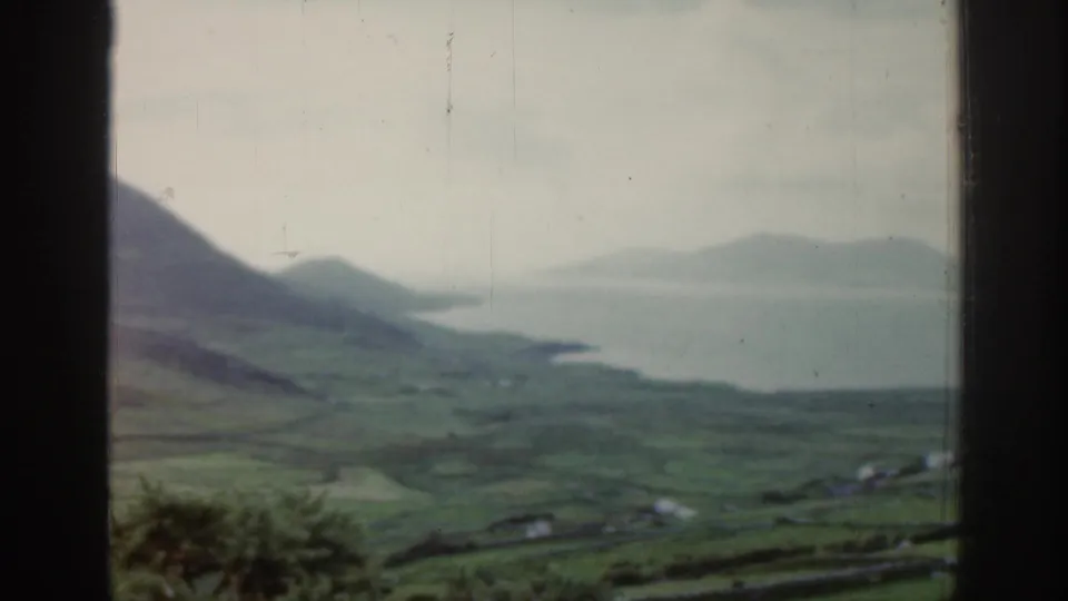 1961 Ireland travel frame looking over green hills toward a coastal bay and hazy distant landforms.