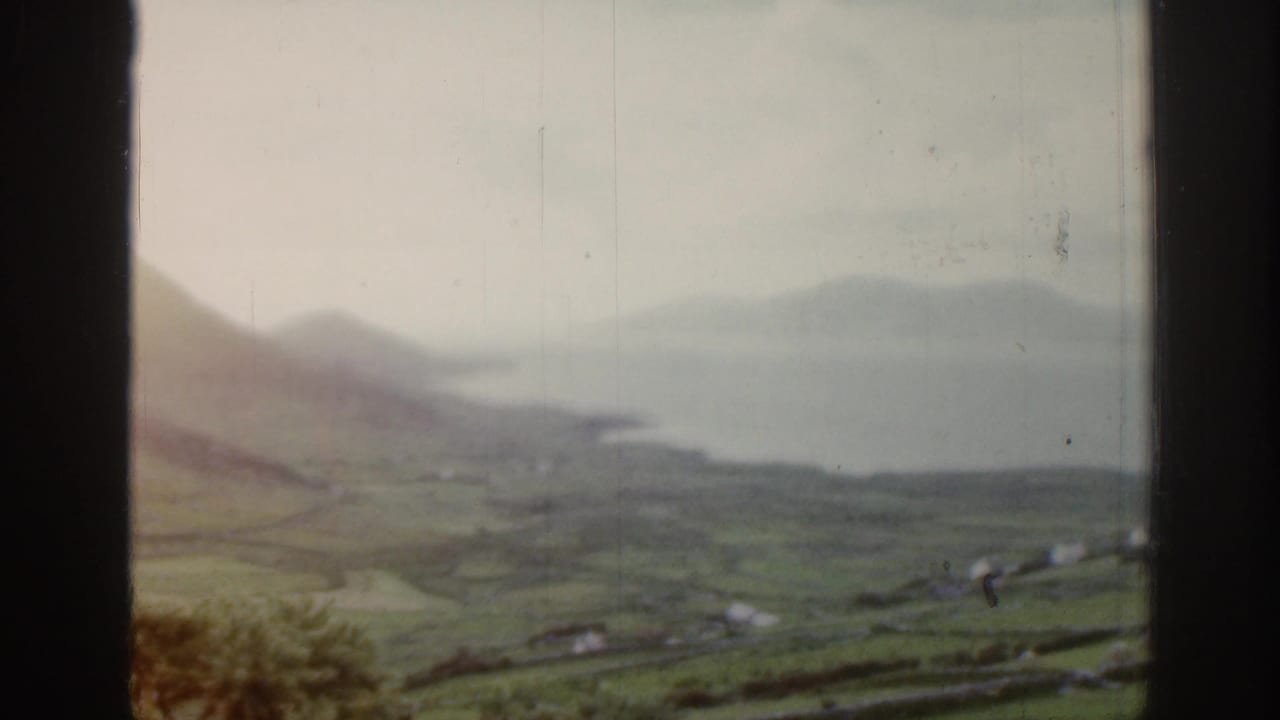 Soft 8mm frame from Ireland showing hillside fields, coastal water, and scattered rural buildings in the distance.