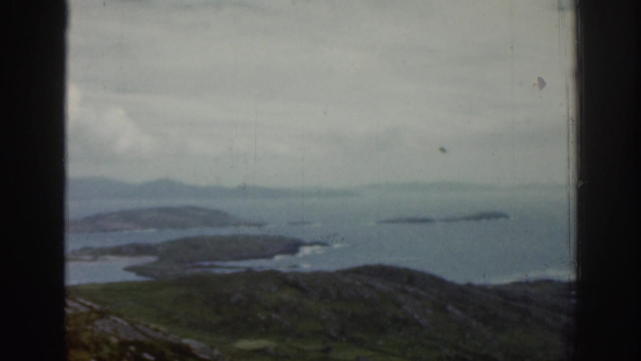 1961 Ireland 8mm frame showing a soft coastal landscape through a window, with green hills, blue-gray water, small islands, and a hazy horizon.