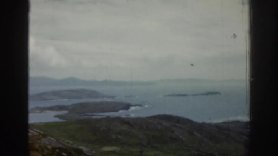 1961 Ireland 8mm frame showing a soft coastal landscape through a window, with green hills, blue-gray water, small islands, and a hazy horizon.