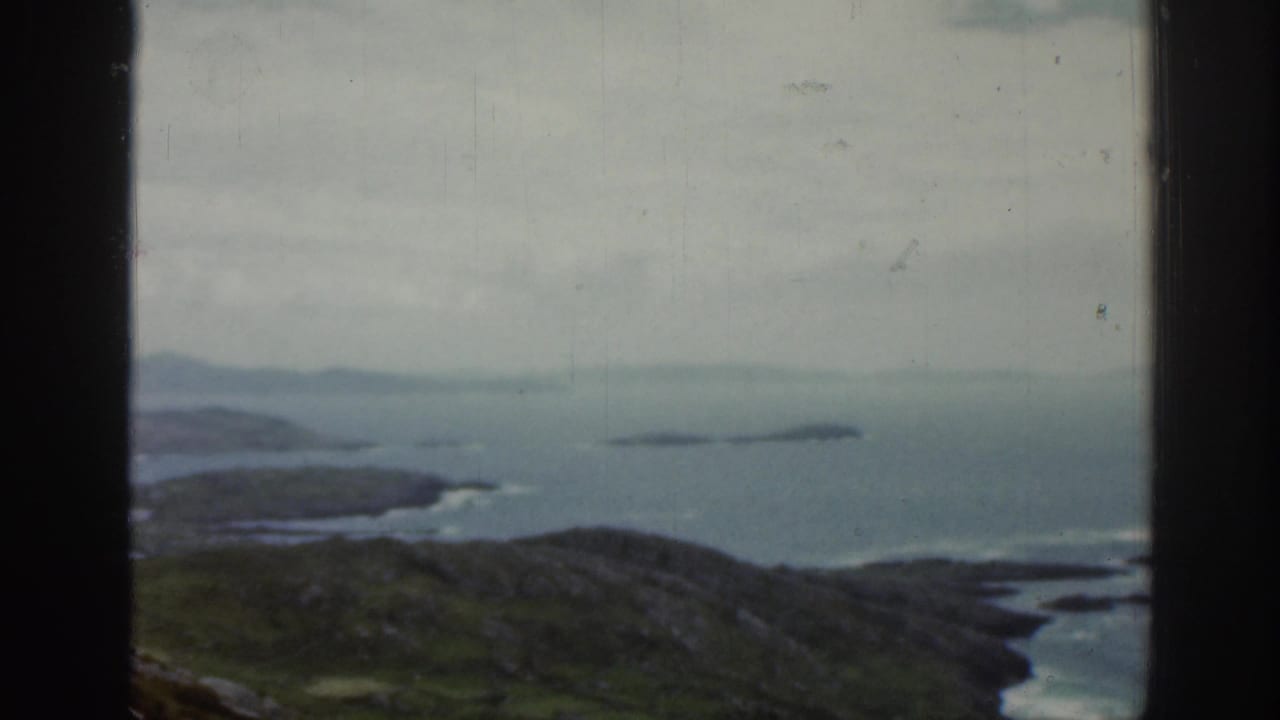 Window-framed seaside landscape with rocky green foreground, calm water, distant islands, and cloudy sky.