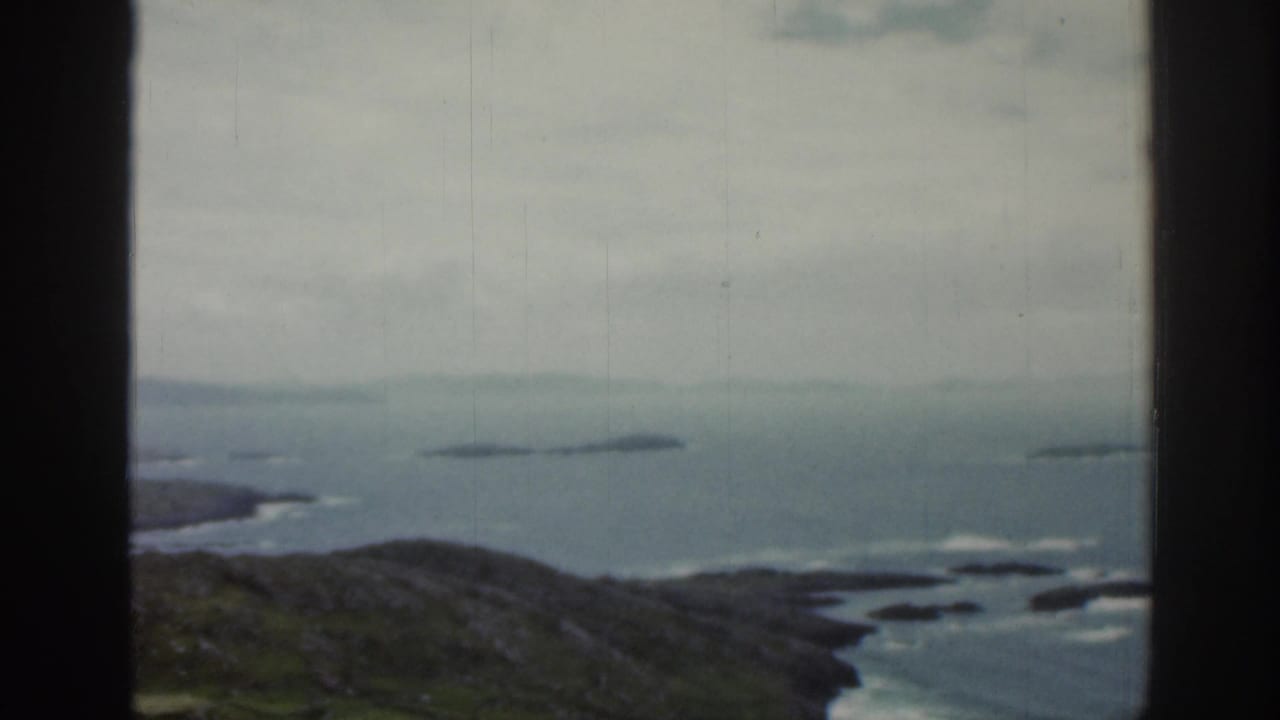 1961 Ireland vacation footage frame looking through a window toward a green headland, calm blue-gray water, small islands, and overcast haze.