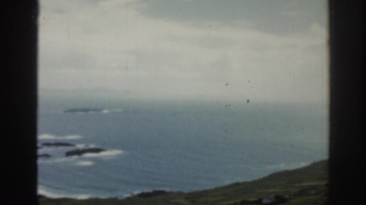 1961 Ireland home-movie frame showing Atlantic coastline, green slopes, and a dark aircraft-window divider.
