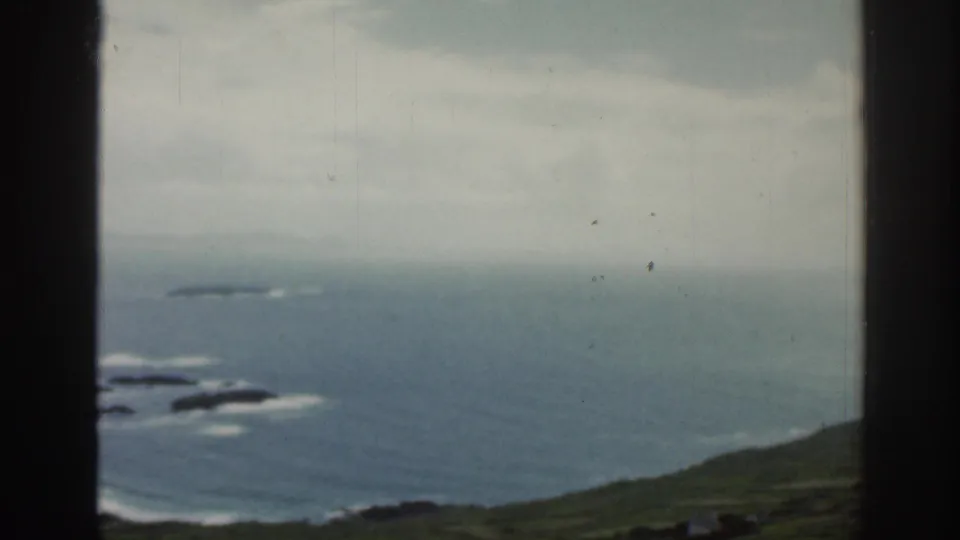 1961 Ireland home-movie frame showing Atlantic coastline, green slopes, and a dark aircraft-window divider.