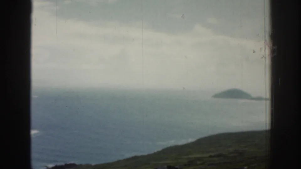 1961 Ireland 8mm frame looking over green coastal terrain toward a small offshore island and hazy Atlantic water.