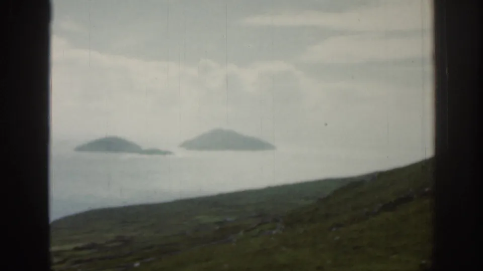 1961 Ireland aerial frame showing two low islands offshore, a green hillside below, and a cloudy sky through the window.
