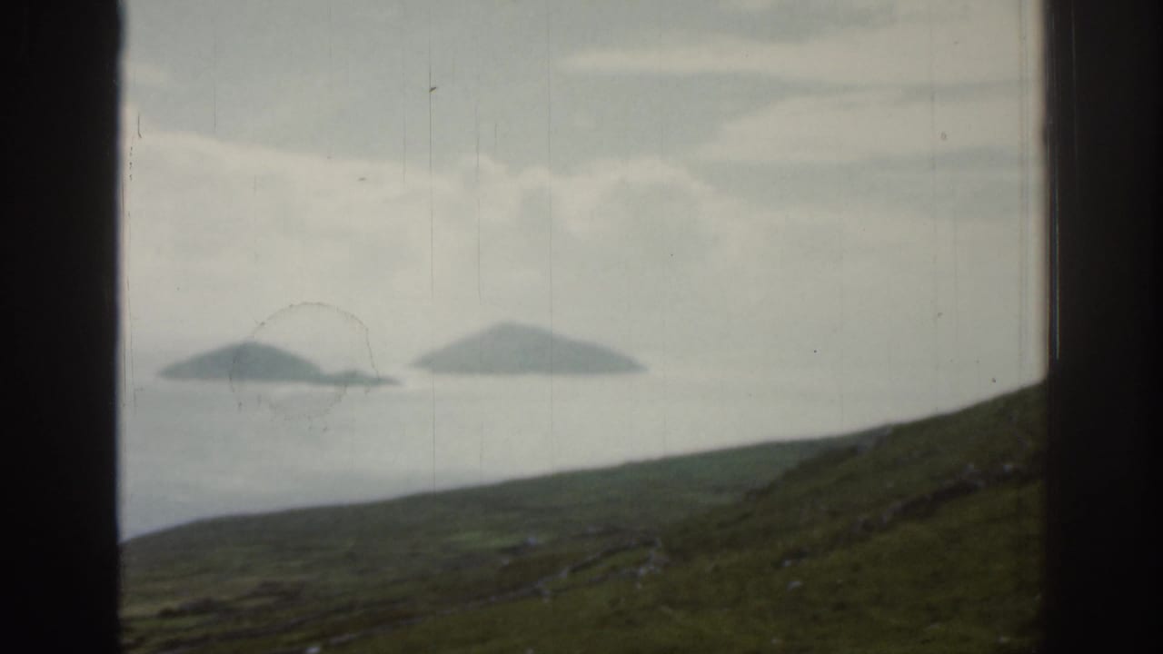 Distant islands and sea seen through a dark window frame above a green hillside.