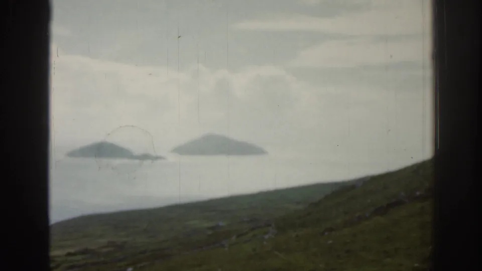 1961 Ireland coastal view through a dark window frame, with distant islands, misty water, and a sloping green hillside.