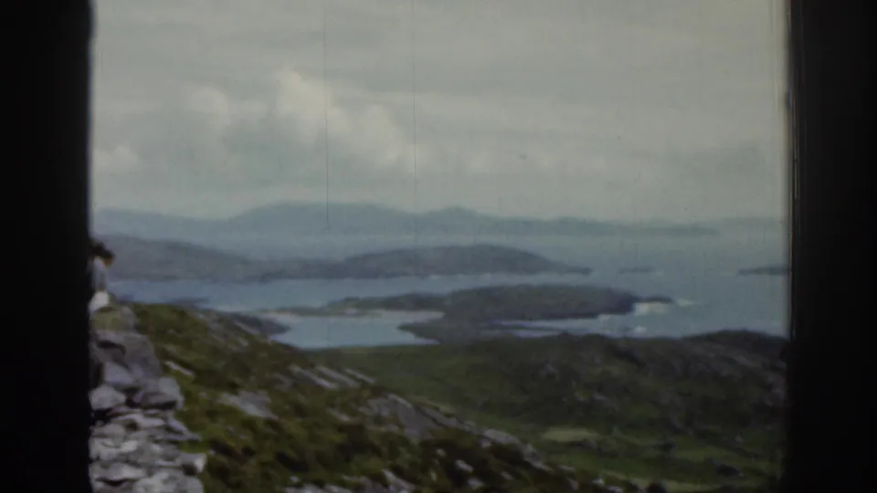 1961 Ireland home-movie frame of rocky green ground above broad water, islands, and distant hazy hills.