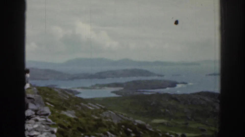 1961 Ireland coastal panorama with rocky foreground, blue-gray water, distant hills, and a partial person near the frame edge.