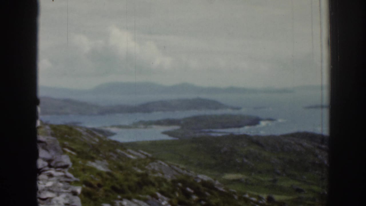 Rocky green hills and coastal water seen through a dark window frame.