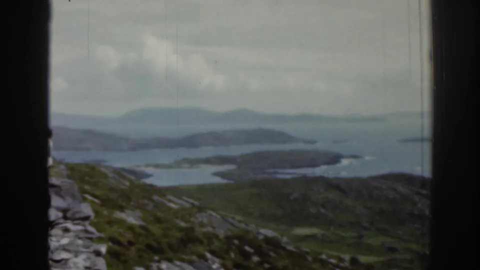 1961 Ireland home-movie frame showing rocky grass-covered hills sloping toward coastal water, divided by a dark window frame under cloudy sky.