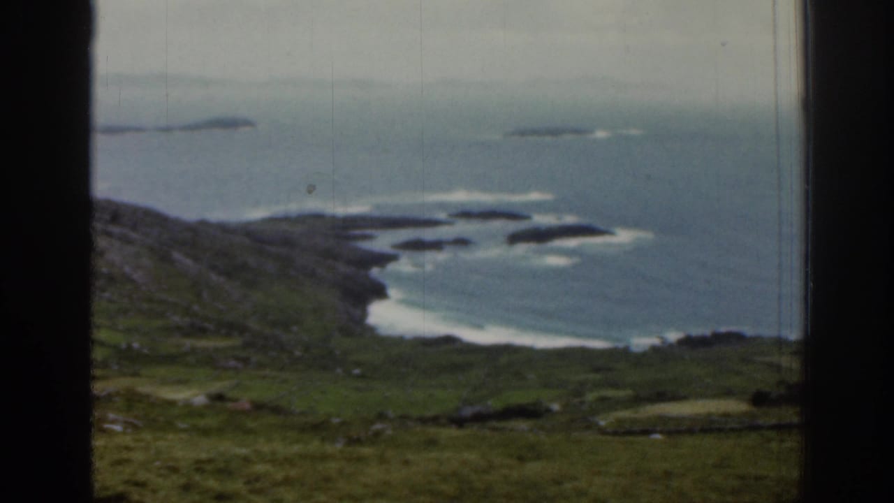 1961 Ireland coastal view with grass-covered terrain dropping toward a rocky shoreline, pale surf, small islands, and a dark vertical window divider.