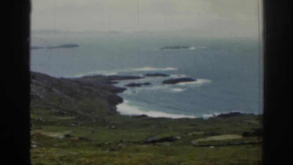 1961 Ireland frame showing a grassy coastal slope above blue water, white surf along the rocks, and small offshore islands beneath overcast light.