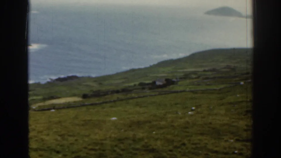 1961 Ireland coastal hillside view with open grassland, stone field boundaries, distant ocean surf, and a small island on the horizon.