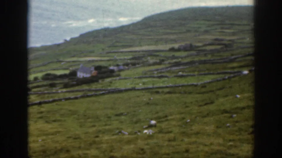1961 Ireland home-movie view of sheep grazing below stone-walled hillside fields and a cluster of small cottages.