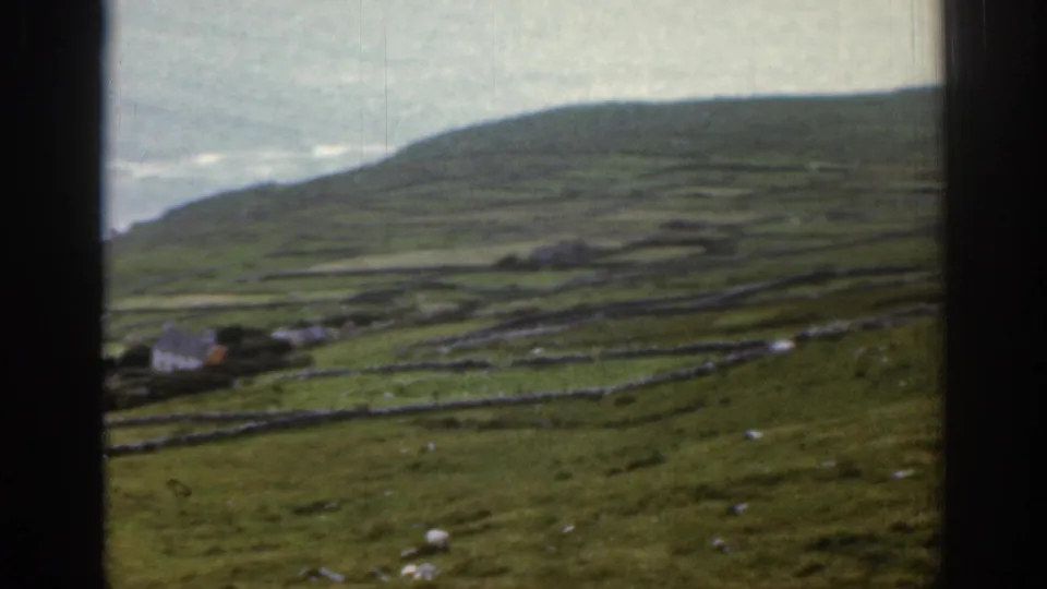 1961 Ireland frame showing rolling coastal farmland, stone field walls, small cottages, sheep, and a partial ocean view.