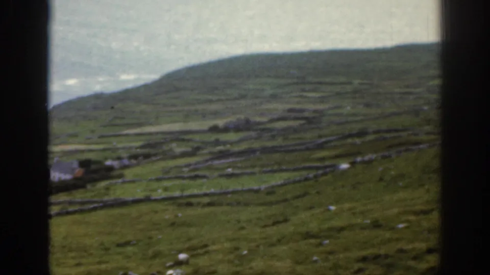 1961 Ireland 8mm view of a grassy coastal hillside divided by stone walls, with small white buildings and sea beyond.