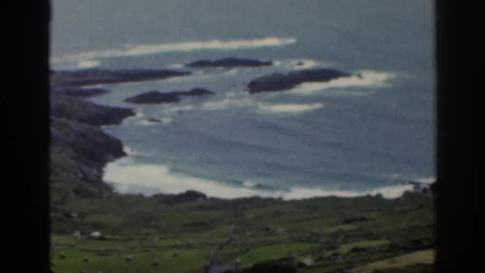 1961 Ireland 8mm frame of a bay with offshore islands, breaking surf, green fields, and stone wall divisions near the coast.