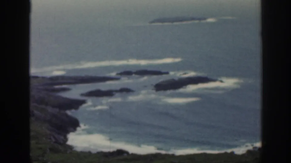 1961 Ireland 8mm coastal frame with a blue bay, dark rocky outcrops, small islands, and white surf patterns offshore.