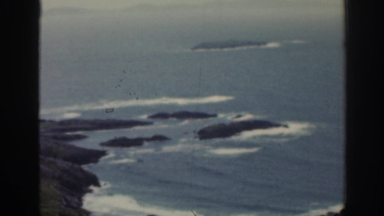 1961 Ireland 8mm frame showing hazy ocean, rocky outcrops, and surf viewed through a dark window mullion.